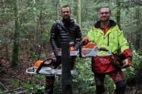 Zwei Männer stehen in jungem Wald hinter zwei Motorsägen die sie kreativ in einen umgeschnittenen Baum fest gesägt haben.